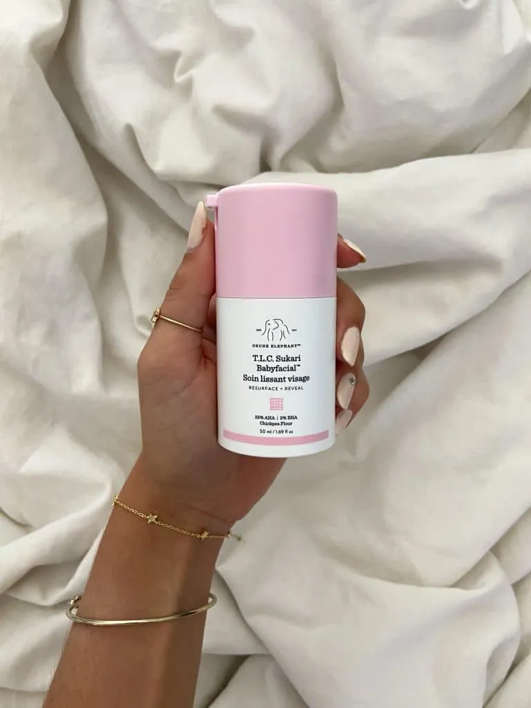 Hand holding the pink Drunk Elephant T.L.C. Sukari Babyfacial bottle over white sheets
