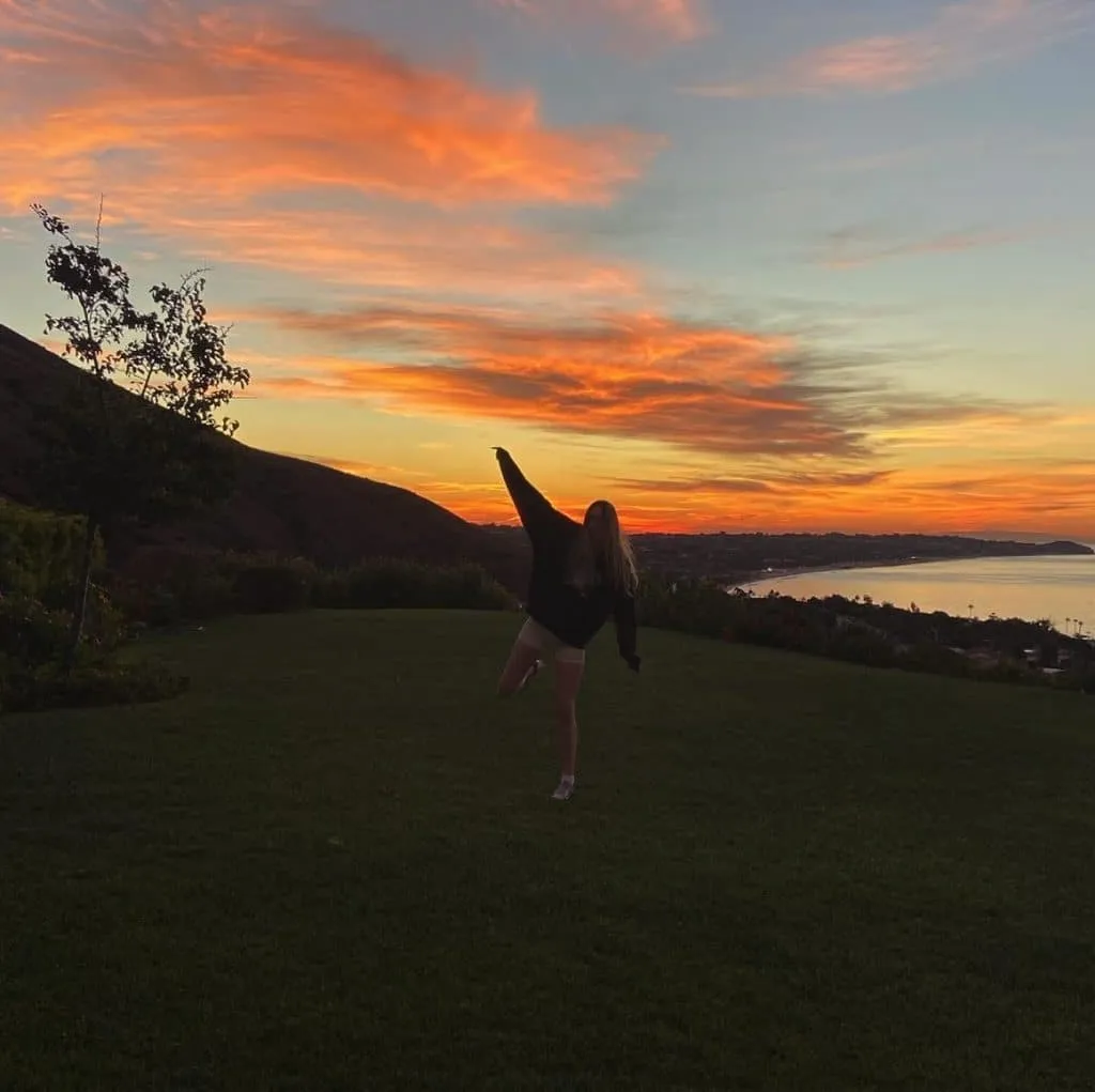 Sky in an oversized dark sweatshirt and light bike shorts posing on a hill at sunset over the coast