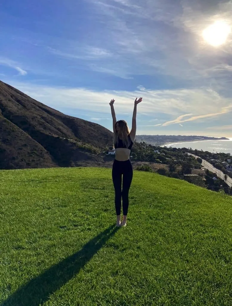 Sky in a black Set Active sports bra and leggings barefoot on a grassy hill with arms raised toward the sky