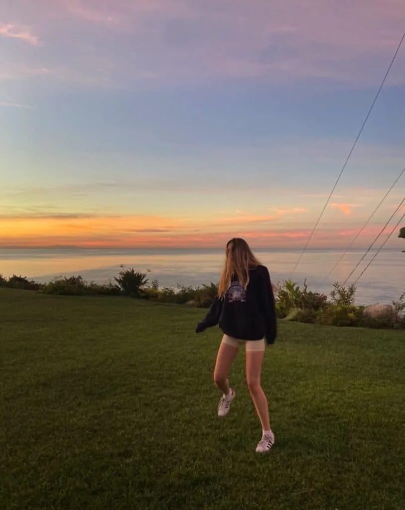 Sky in a dark sweatshirt, light athletic shorts and Adidas sneakers on a lawn overlooking the ocean at golden hour