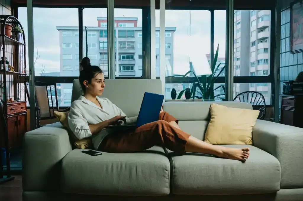 Woman in a white shirt working on a blue laptop on a light gray sofa in a city apartment with a tall bookshelf