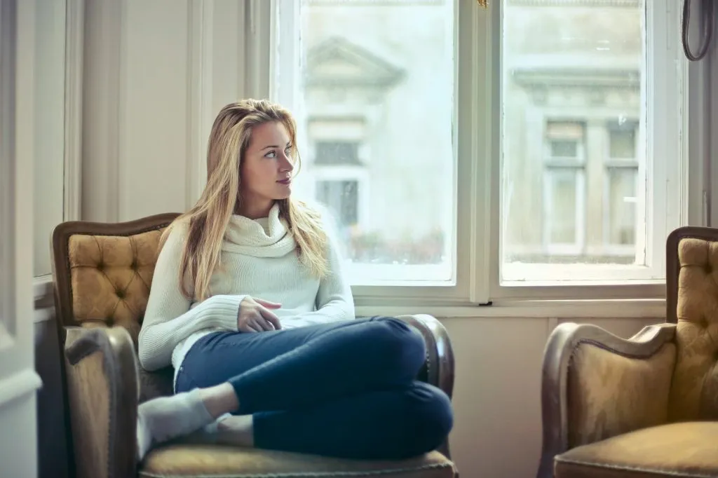 Woman with long blonde hair in a cream sweater sitting in a mustard-yellow armchair by a window