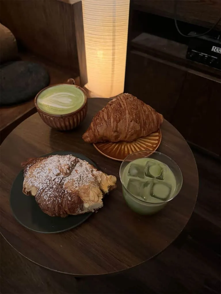 Hot matcha latte, an iced matcha and two croissants on a dark wood table under a paper lamp at 915 Dupont