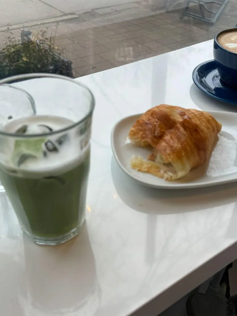 Iced matcha latte in a glass next to a croissant and a coffee by the window at Buno Coffee
