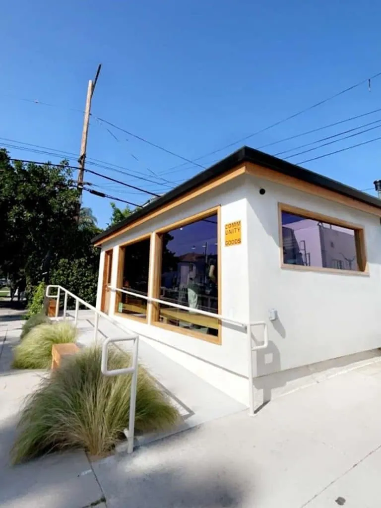 White stucco Community Goods cafe exterior with wood-trimmed windows, a yellow sign and fountain grass out front