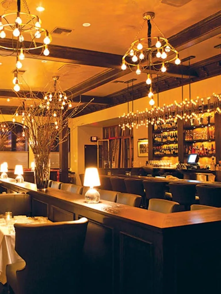 Warm amber-lit Crossroads Kitchen bar with exposed-bulb chandeliers, dark wood and a stocked back bar