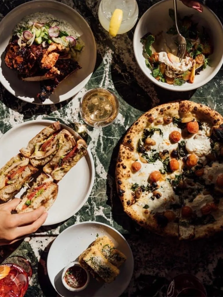 Overhead spread at Great White: pizza with burrata and tomatoes, sandwiches, salad, toasted bread and cocktails on green marble