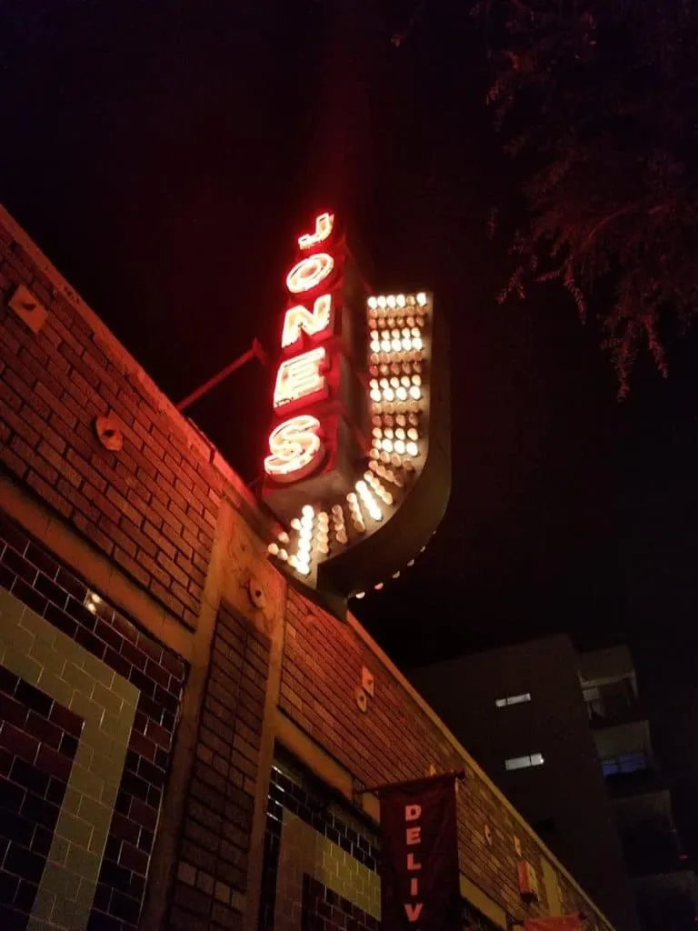 Nighttime neon Jones marquee sign with red letters and rows of warm bulbs on a brick building