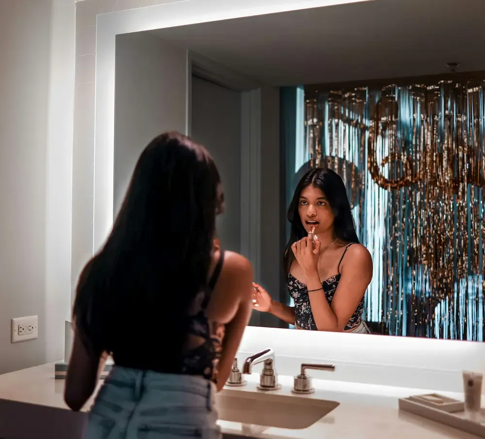 Woman applying lipstick at a large bathroom mirror above a white sink, reflecting a chandelier
