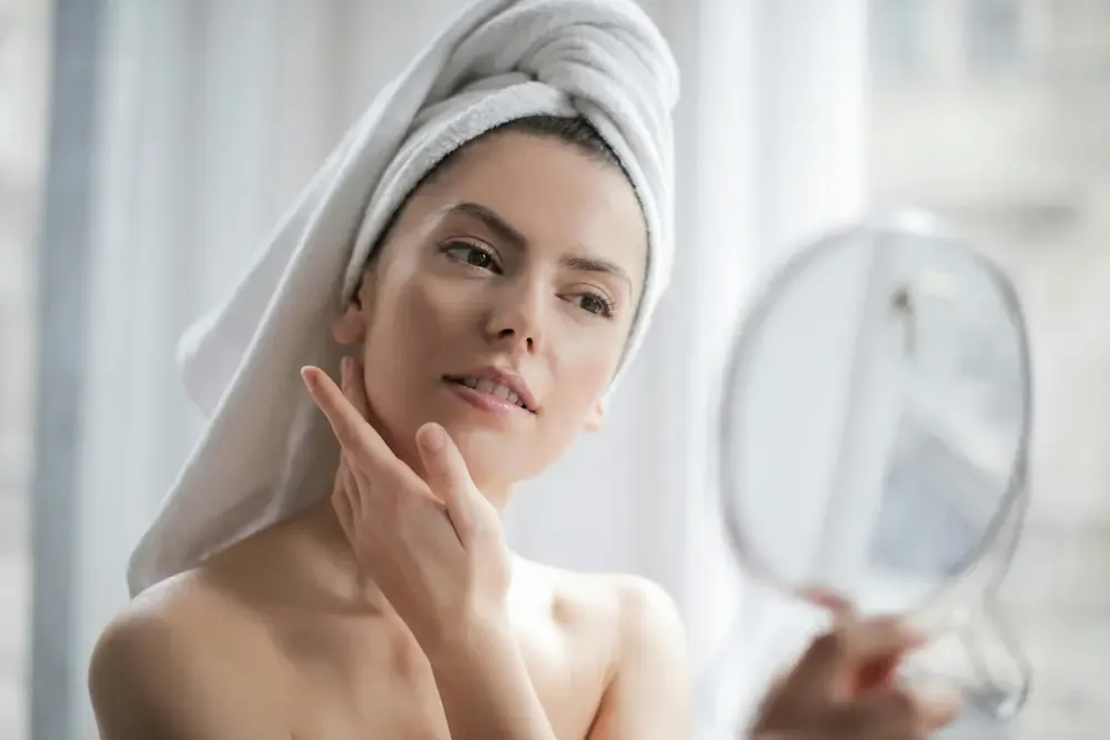 Woman in a white hair towel admiring her smooth, dewy skin in a round hand mirror