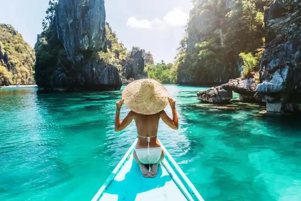 Woman in a white crochet bikini and oversized straw hat on a blue boat looking out at turquoise water and green cliffs