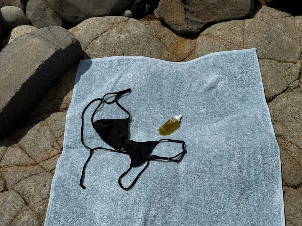 Black string bikini top and a clear dropper bottle of golden Aiya hair oil on a light blue terry towel spread over rocks
