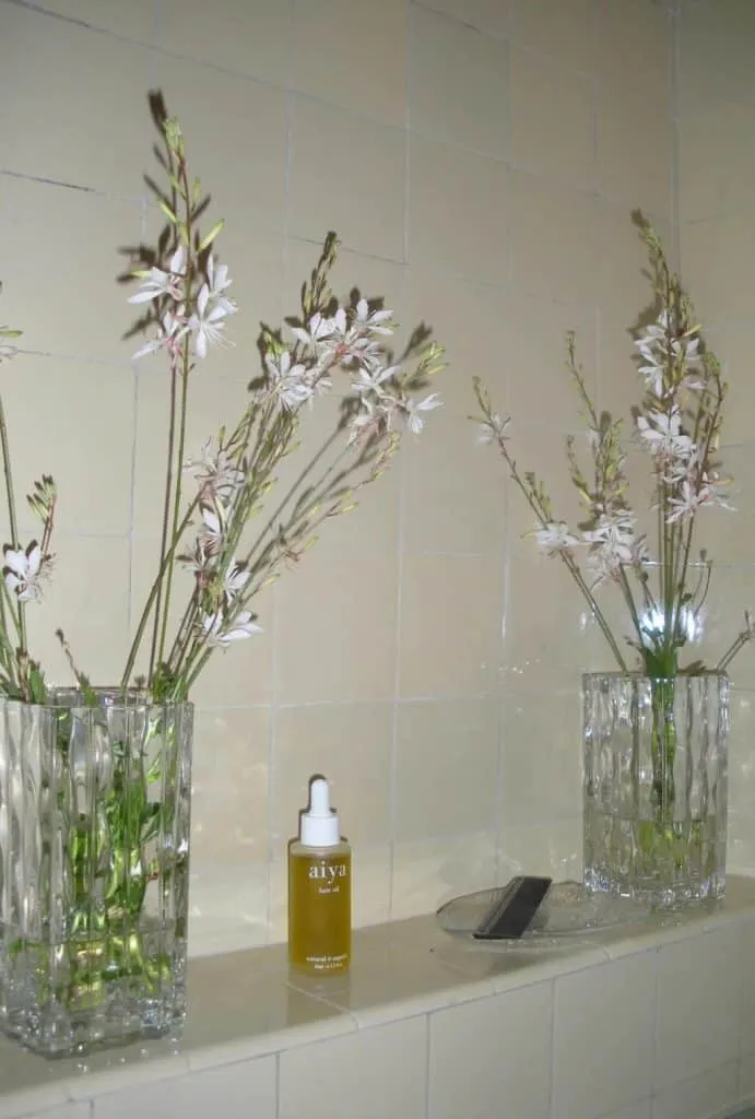 Clear dropper bottle of golden Aiya hair oil on a beige tiled ledge between two glass vases of white wildflowers and a dark comb