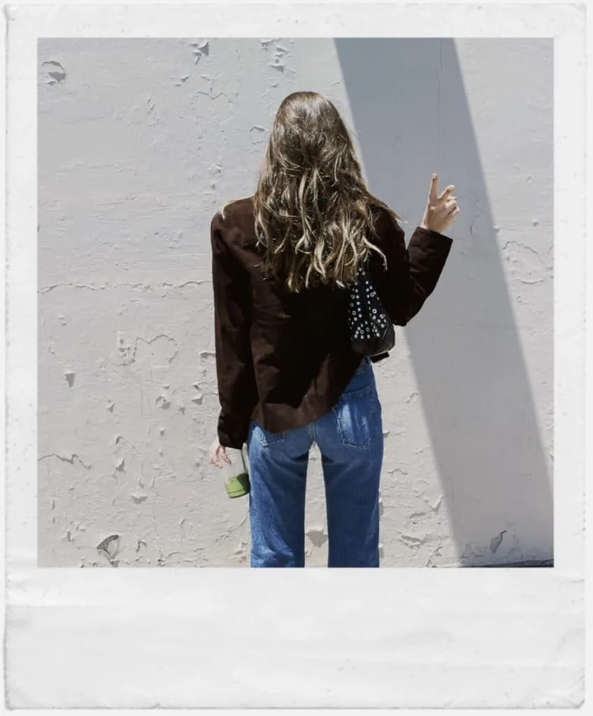 Polaroid-style back view of Sky in a mocha suede jacket and jeans flashing a peace sign with an iced matcha in hand