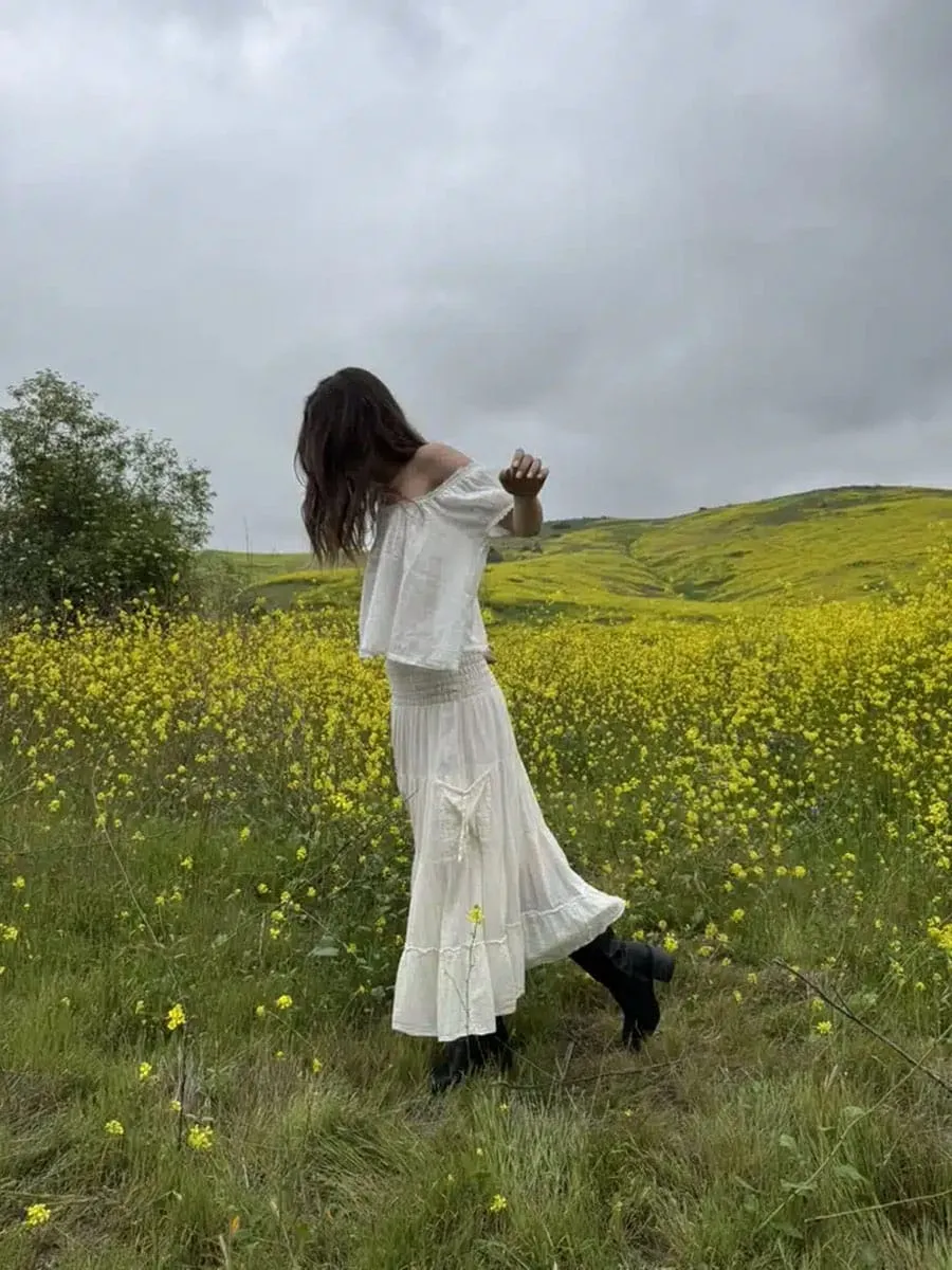 Sky in a white DÔEN Frederica Top, a long cream skirt and knee-high black boots in a yellow wildflower field under grey skies