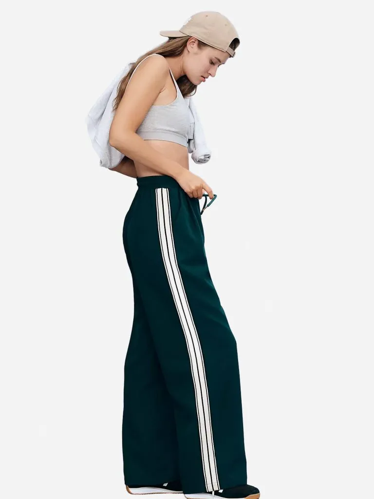 Model in dark green track pants with white side stripes, a gray sports bra and a beige cap