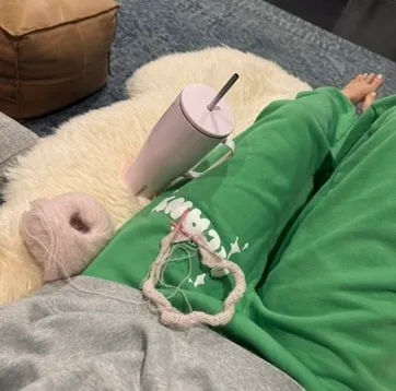 Corkcicle tumbler on a sheepskin rug beside a knitting project and pink yarn, legs in green sweatpants on the couch