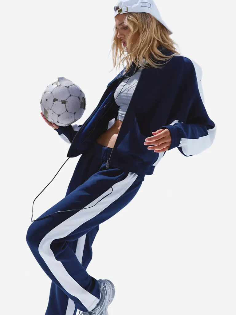 Woman in a navy tracksuit with white side stripes, grey cropped tank, white cap worn backward, balancing a soccer ball