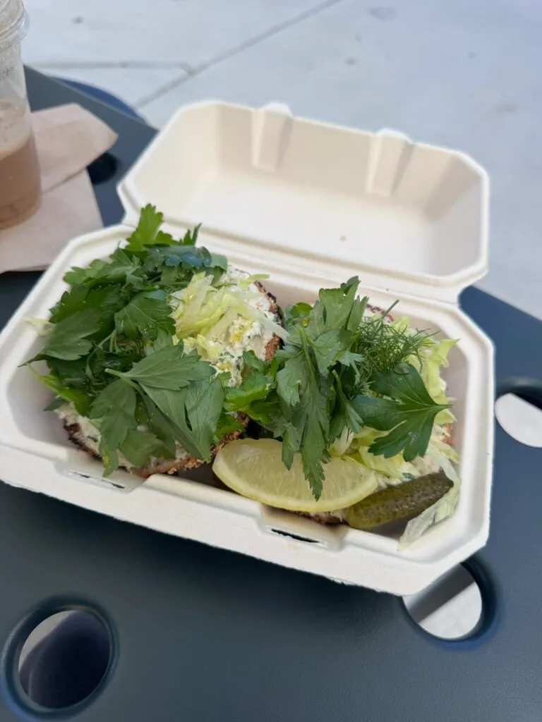 Open-faced bagels piled with fresh herbs, a lemon slice and a pickle in a to-go box from Sour Seed Bagels