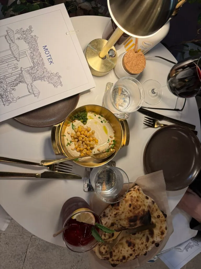Overhead shot of hummus topped with chickpeas in a gold pot, pita, a red cocktail and wine on a white table at Motek South Beach
