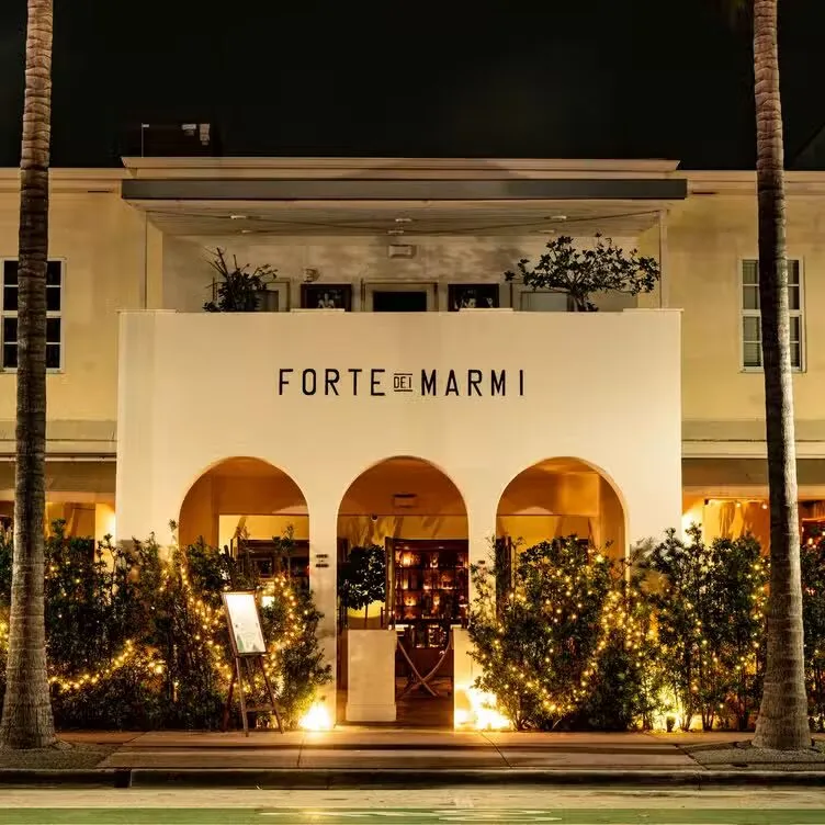 Forte Dei Marmi restaurant in Miami Beach at night, a white arched entrance lined with twinkle-lit hedges and palm trees